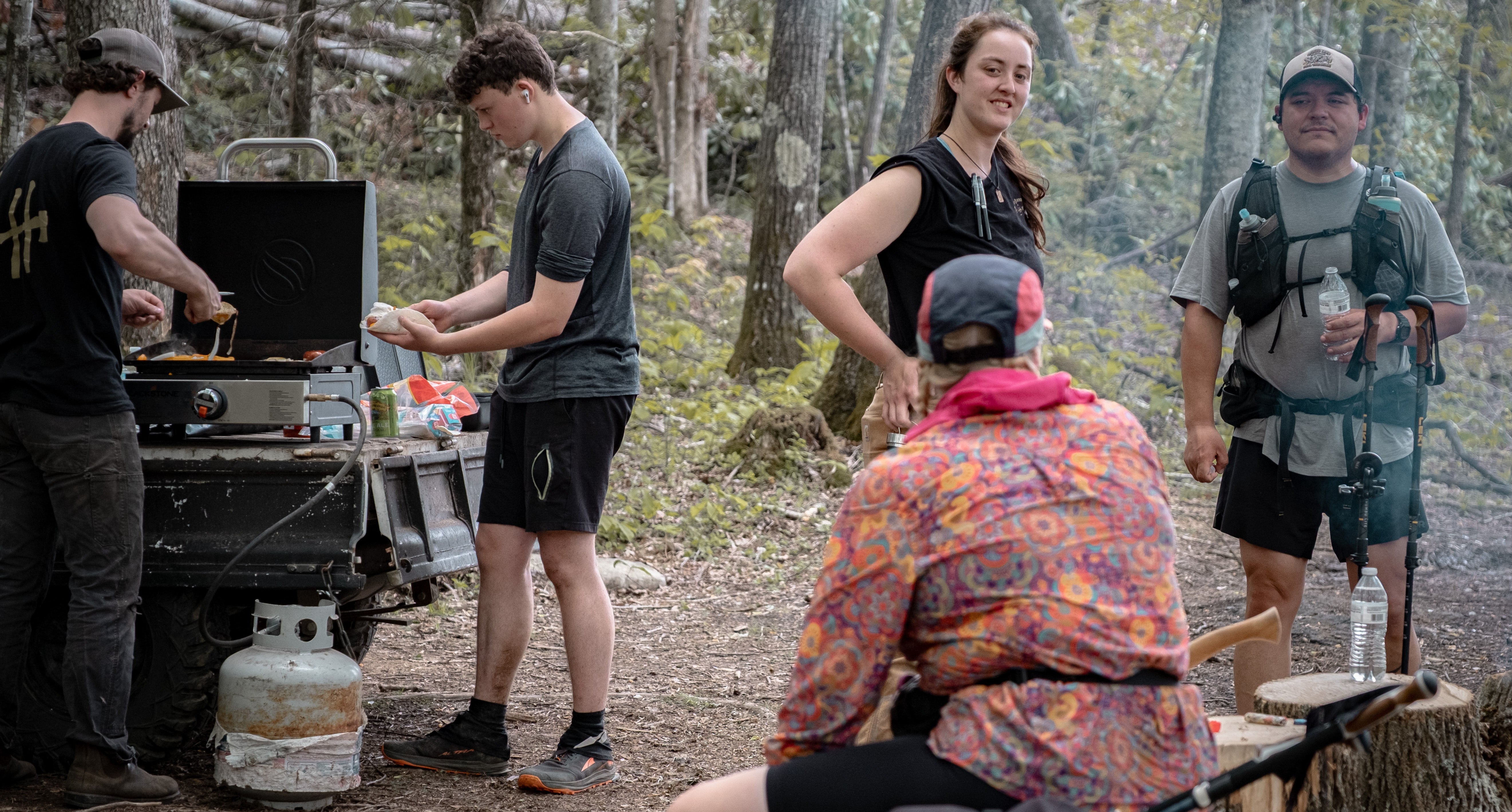 Group of people camping in a forest, preparing food and enjoying outdoor activities.