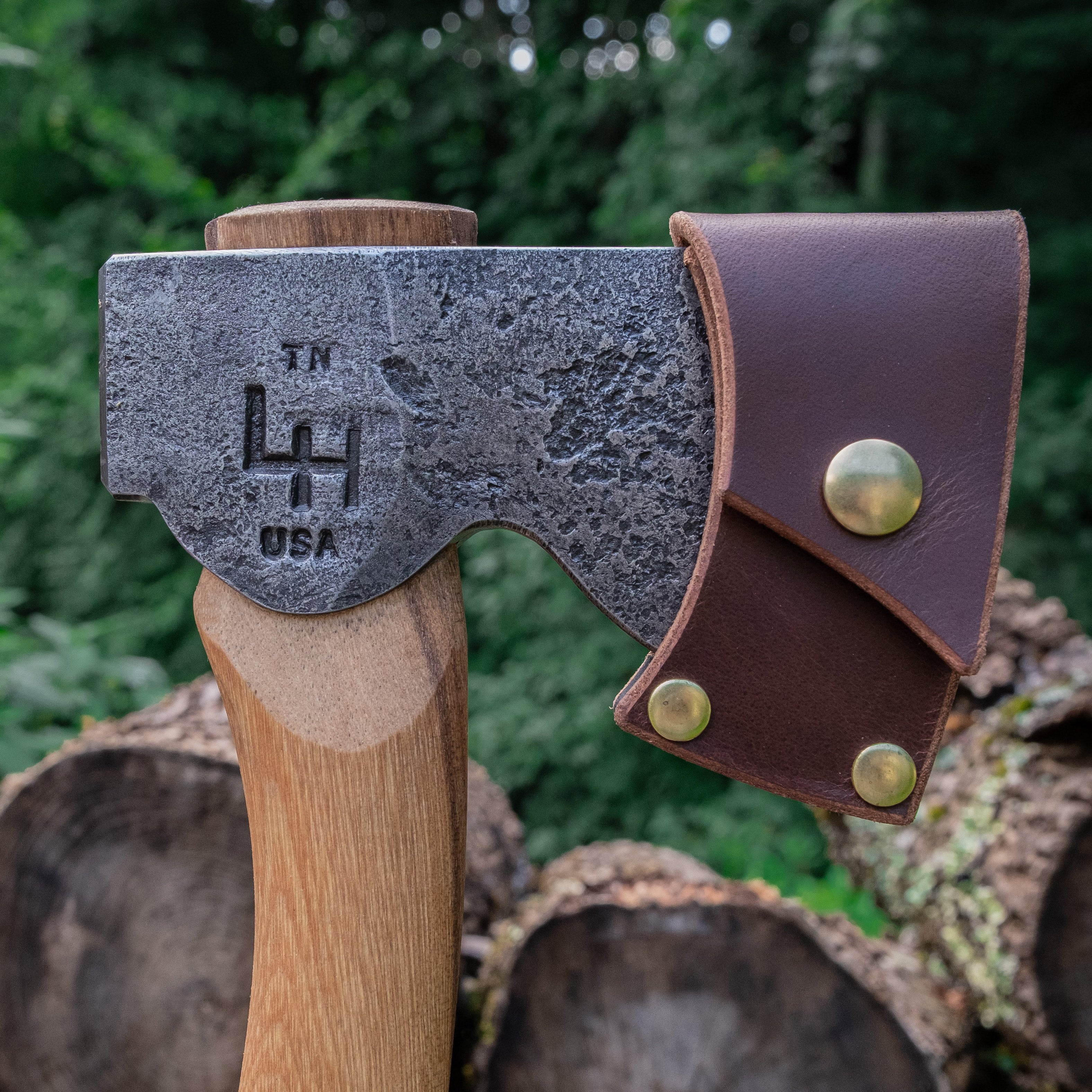 Axes with leather sheaths on a background of stacked logs and greenery