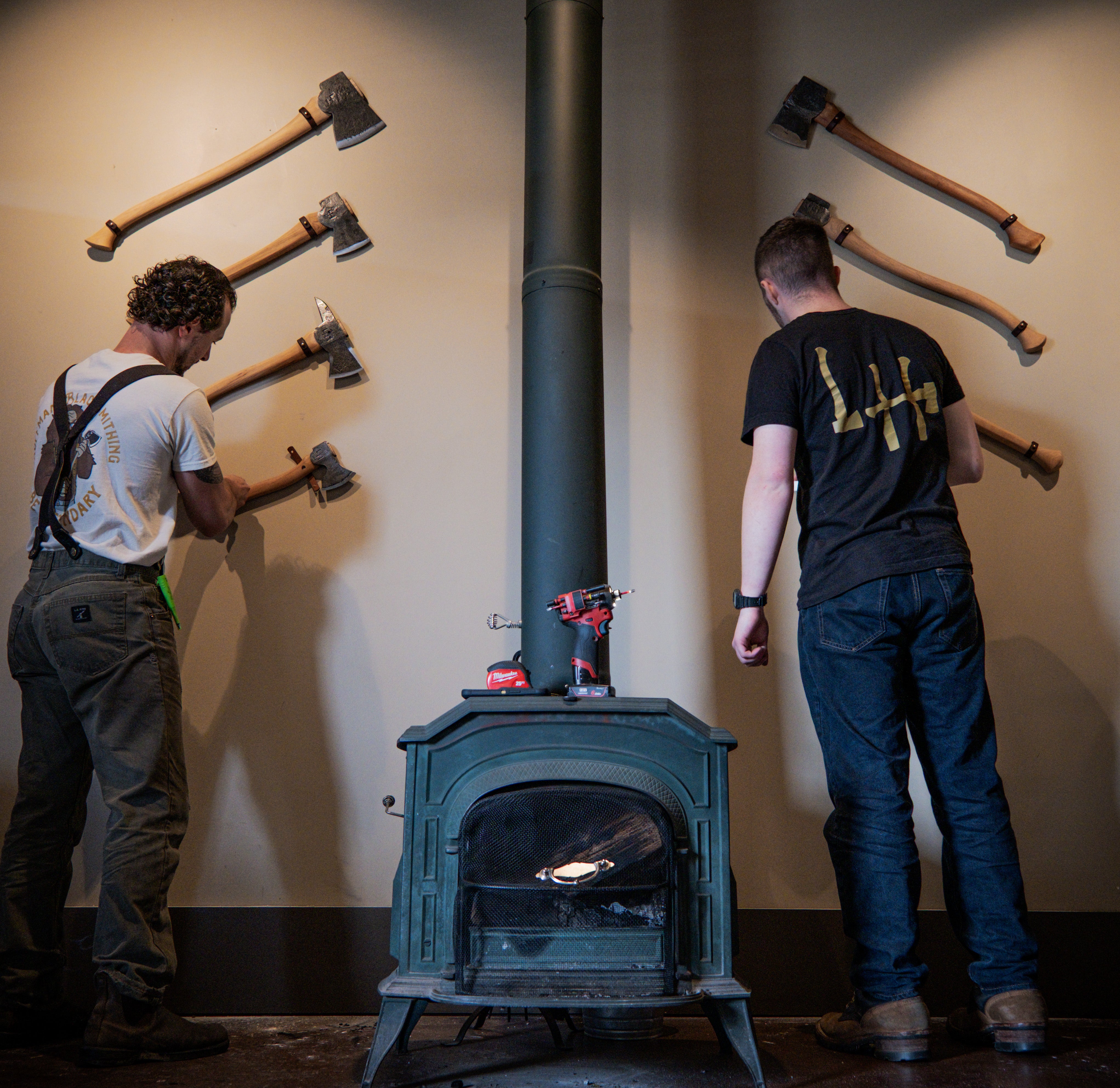 Two men working on a wood stove with axes mounted on the wall behind them.